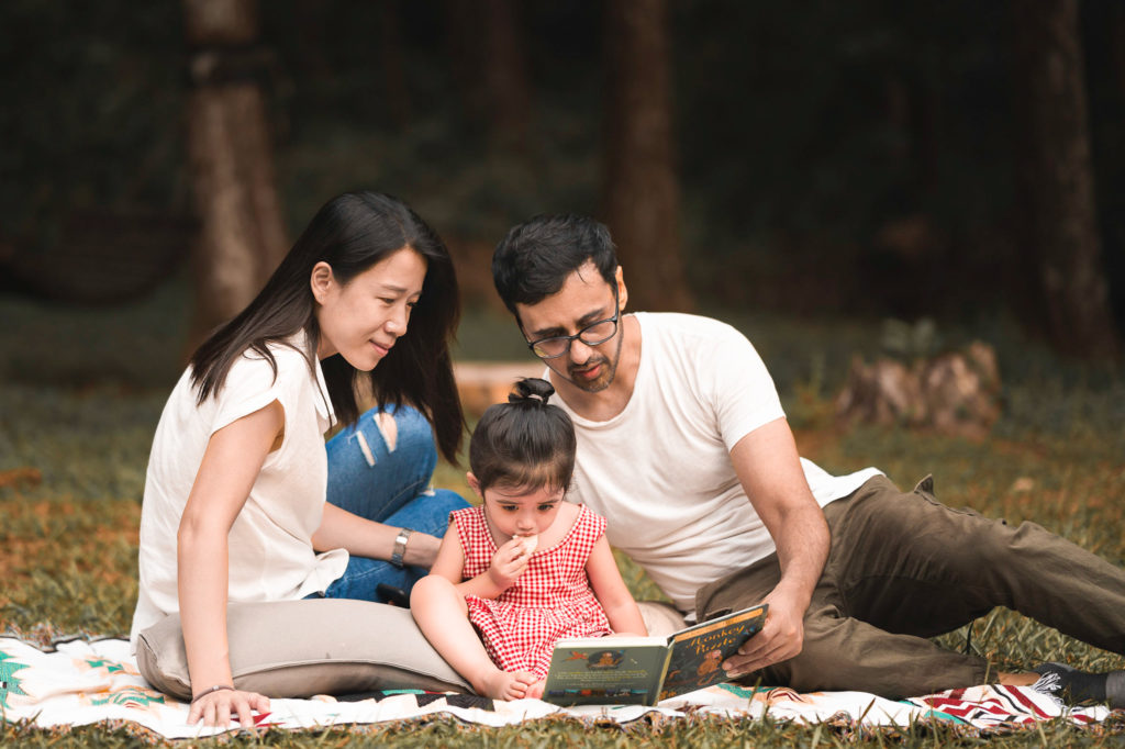 Photo of a family of three sitting on a mat whilst father reads a children's book